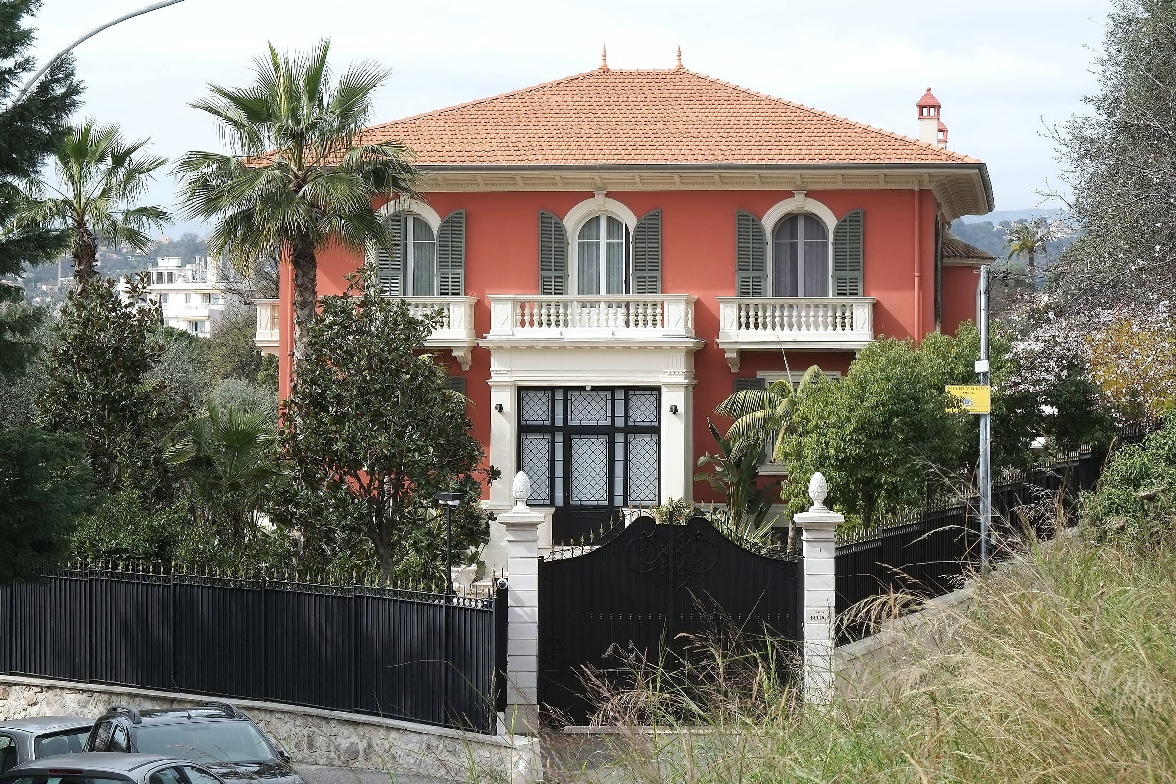 A red villa with white balconies behind a gated garden with palm trees in Cimiez